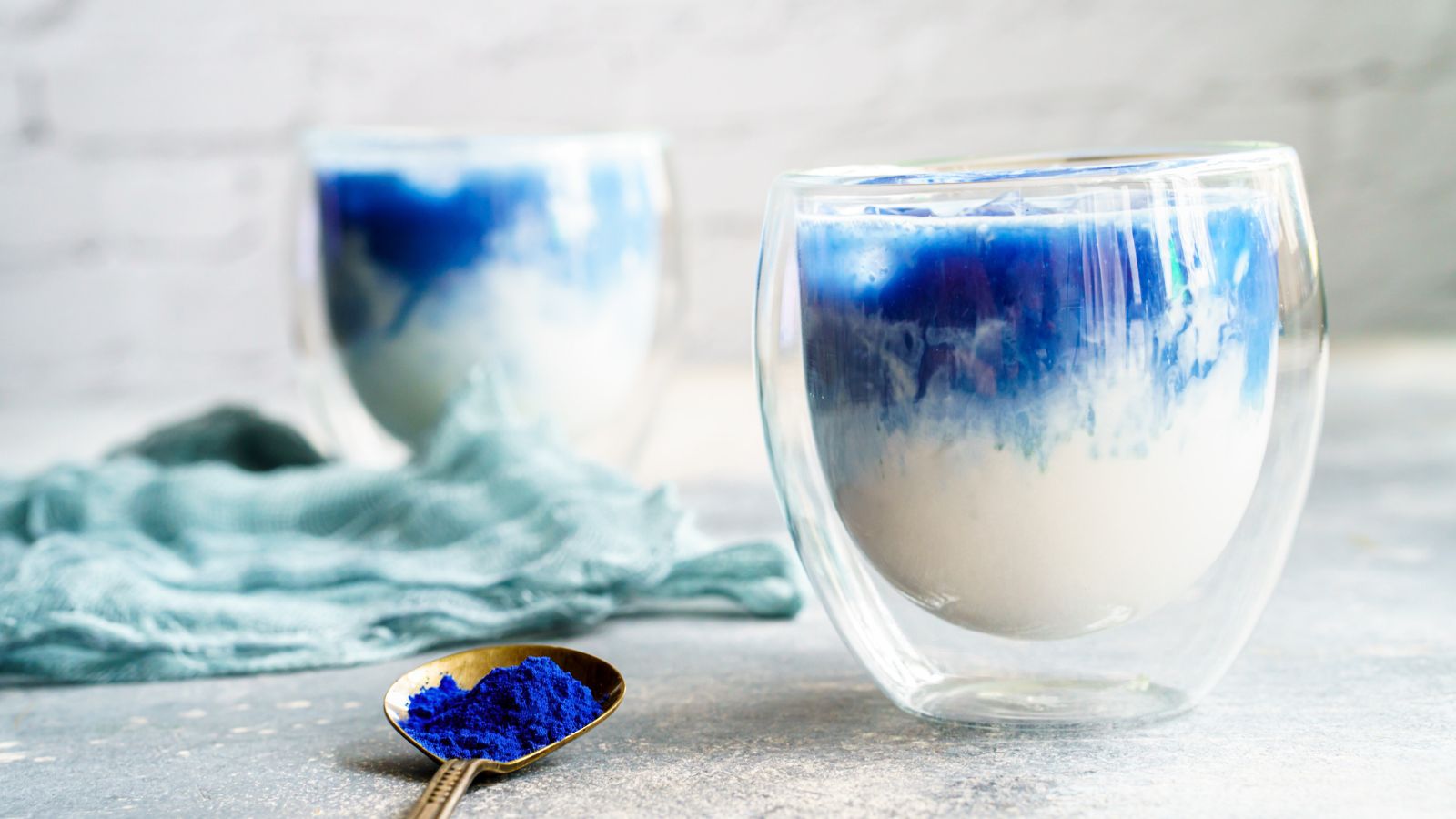 Two glasses of a vibrant blue BlueMAJIK drink, showcasing the antioxidant-rich superfood in a creamy blend, with a spoon of BlueMAJIK powder in the foreground.