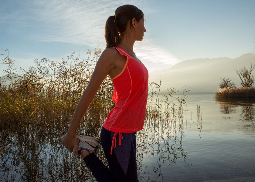 Woman in athletic attire stretching by a serene lakeside at sunrise. The backdrop of mountains and calm water highlights a connection to nature and promotes a sense of balance, strength, and vitality.